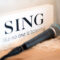 Schild mit der Aufschrift "Sing like no one is listening" mit einem liegenden Mikrofon davor in Szene gesetzt