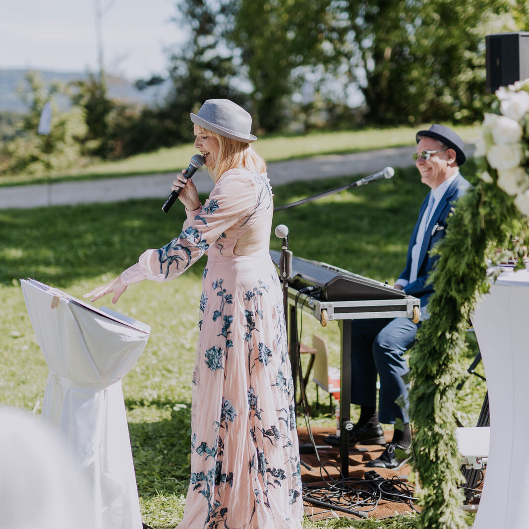 Freie Traurednerin im rosa Kleid mit Blumen verziert und einem Pianisten im Hinterrund hält Ihre Rede im Grünen bei einer freien Trauung.