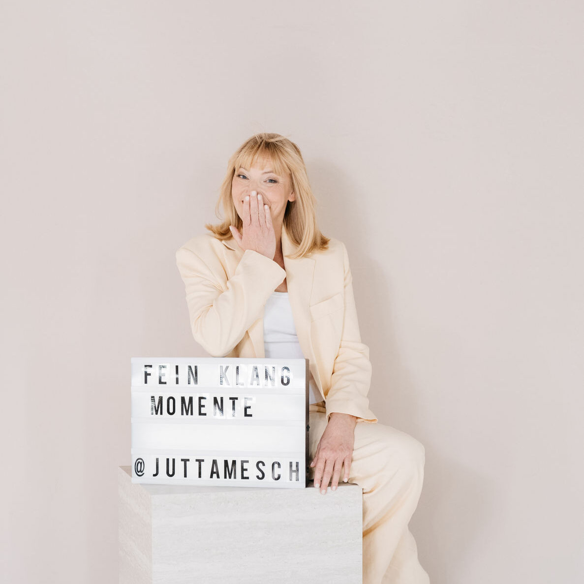 Jutta Mesch sitzt im gelben Hosenanzug mit ihrem Leuchtschild Feinklangmomente.