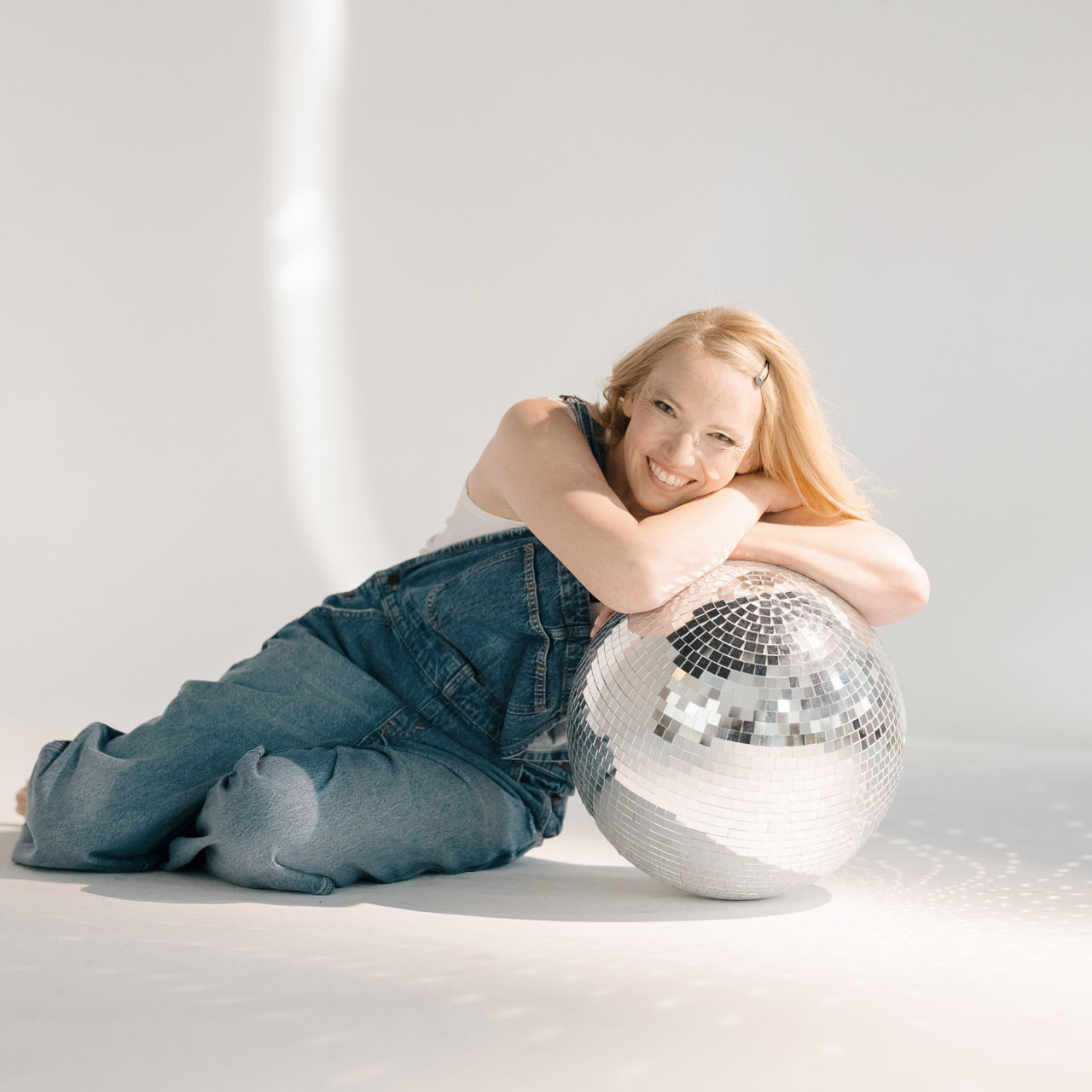 Blondes Mädchen sitzt auf dem Boden mit weißem Hintergrund und lehnt sich auf eine Discokugel