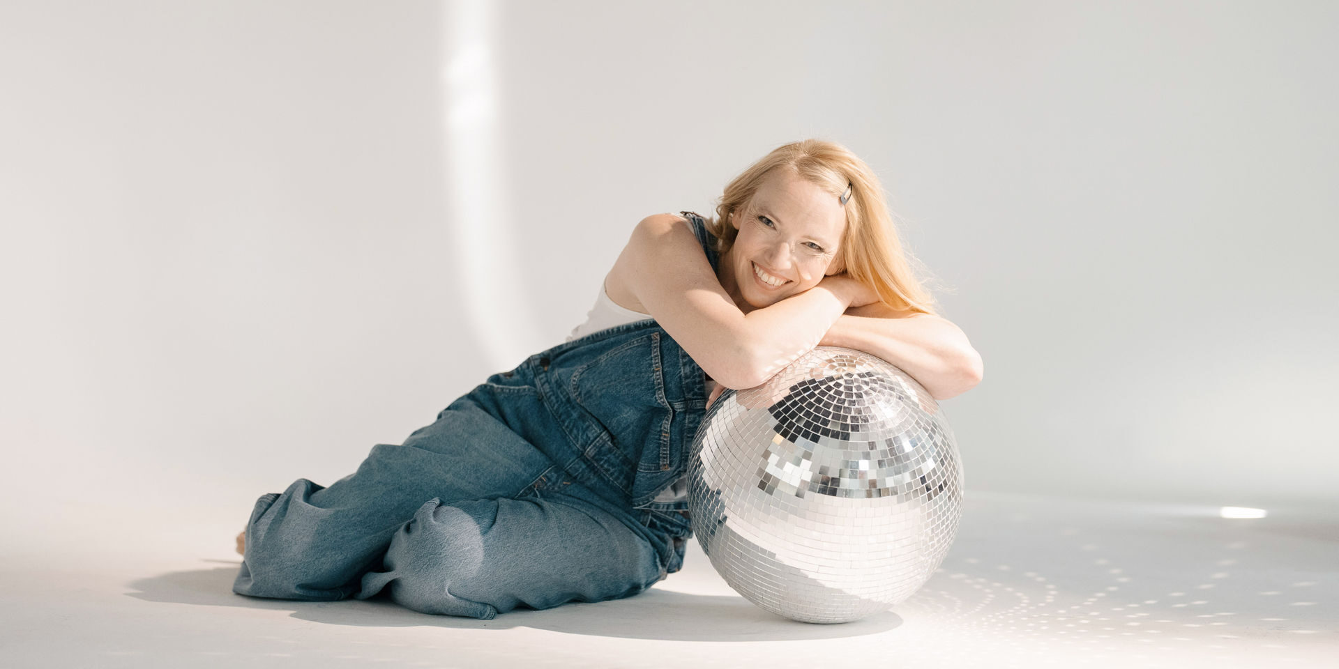 Blondes Mädchen sitzt auf dem Boden mit weißem Hintergrund und lehnt sich auf eine Discokugel