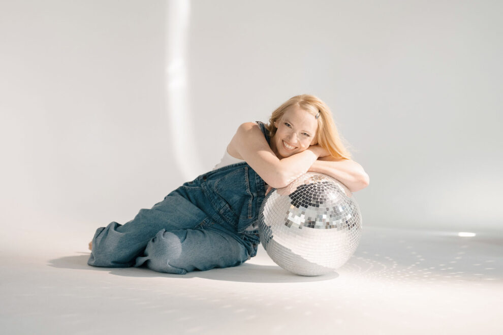 Blondes Mädchen sitzt auf dem Boden mit weißem Hintergrund und lehnt sich auf eine Discokugel